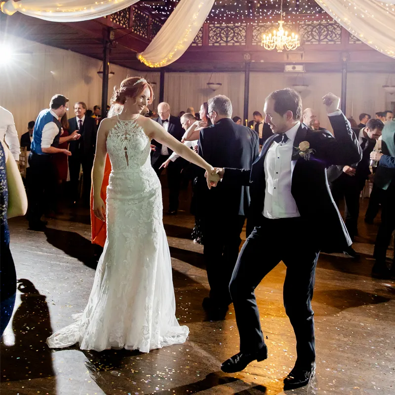 A lively scene captures happy wedding guests dancing joyfully on the dance floor, illuminated by warm evening lights. The atmosphere is filled with upbeat wedding party songs, creating a festive vibe as everyone enjoys the celebration and "wanna dance" together.