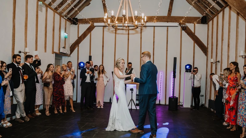 A vibrant wedding reception scene captures a packed dance floor in a rustic barn, where joyful guests are dancing under colorful lights to popular wedding party songs. The atmosphere is lively, filled with the sounds of a DJ Playing and laughter, as everyone enjoys the night long celebration.